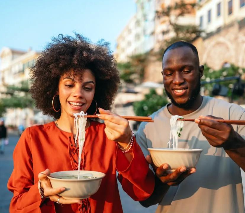 A couple sharing bowls of noodles with chopsticks on a sunny city street