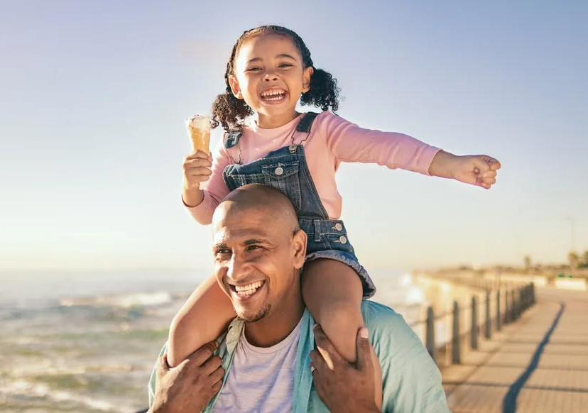 A father with his young daughter on his shoulders, both laughing on a seafront promenade at sunset